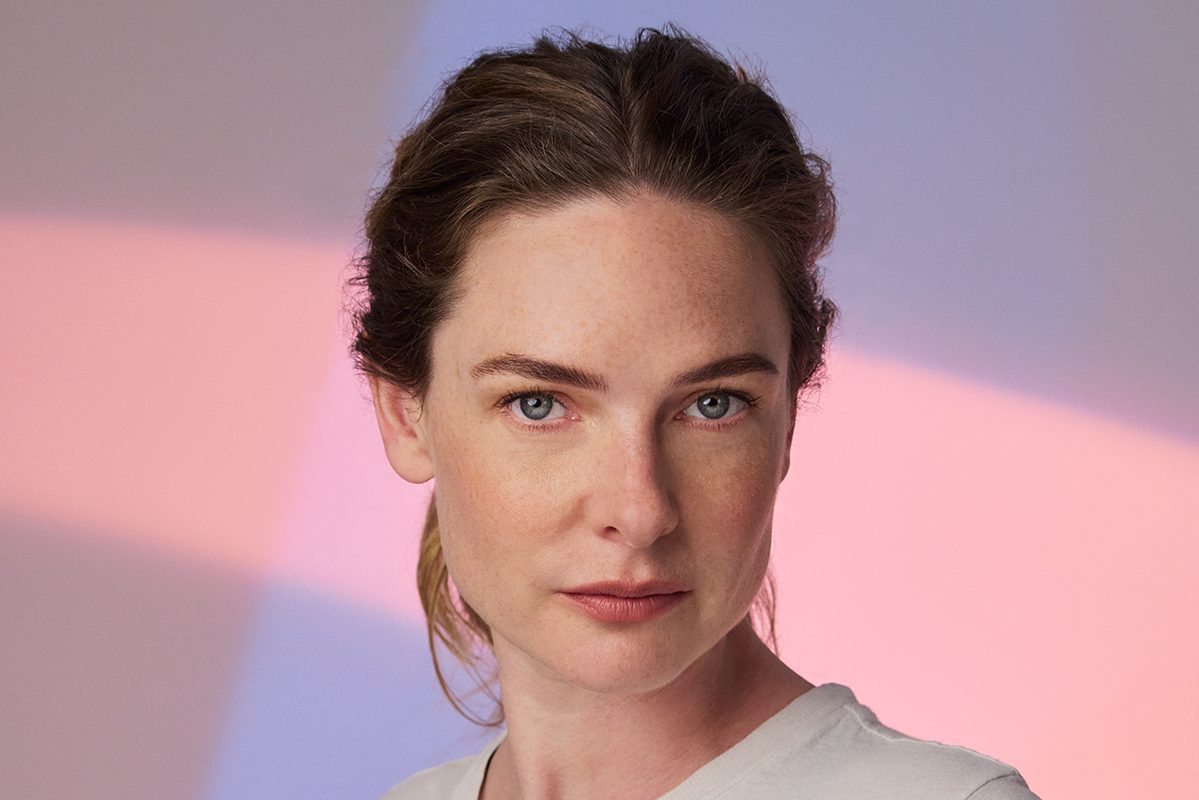 A woman with blue eyes and brown hair, wearing a white t-shirt, is the central figure in a portrait. Her gaze is directed straight at the camera, and she is set against a backdrop of a gradient of pink and blue. The woman's expression is neutral, and her pose is upright, creating a balanced composition